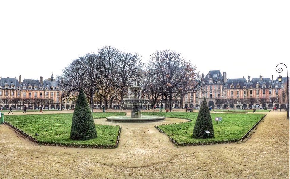 veduta di Place de Vosges (Parigi): una fontana al centro di un giardino con sullo sfondo alberi spogli e circondata da erba tagliata e due siepi di forma conica