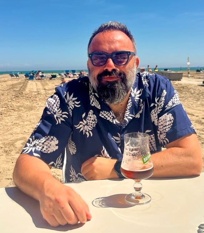 Sorrido indossando occhiali da sole e una camicia blu con ananas a stampe bianche, bicchiere di birra quasi finito, dietro spiaggia, mare e cielo azzurro 