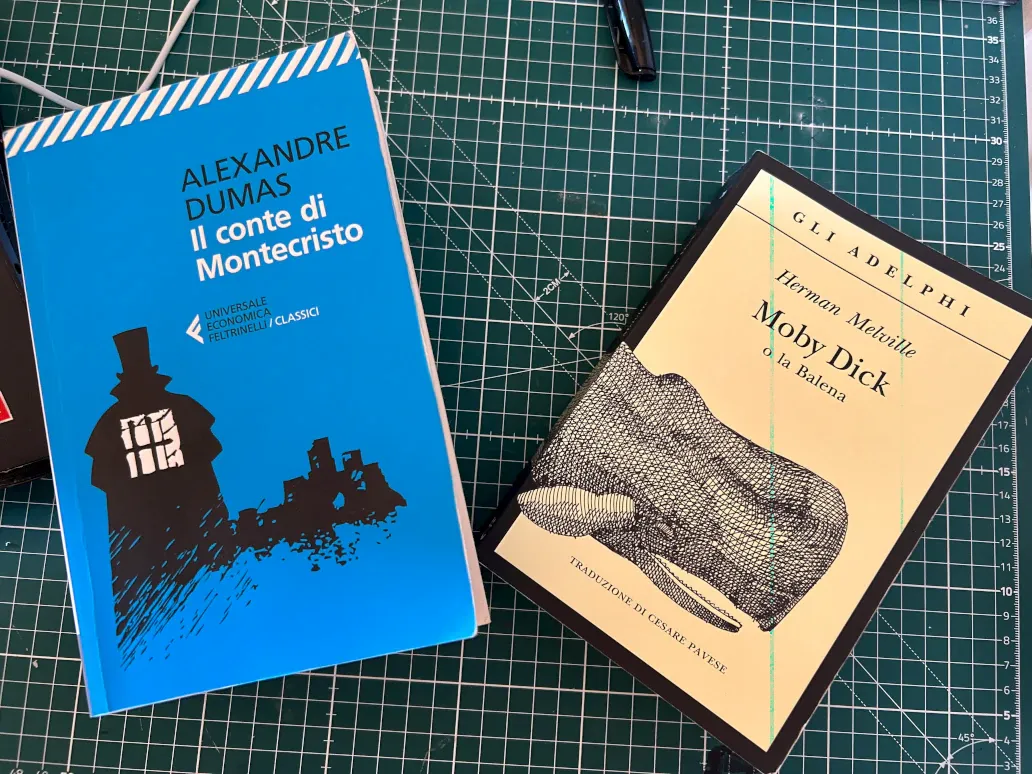 Copertina dei libri del Conte di Montecristo edizione Feltrinelli, sfondo celeste e scritte in bianco e disegno in nero e copertina di Moby Dick edizioni Adelphi, cornice e disegno neri